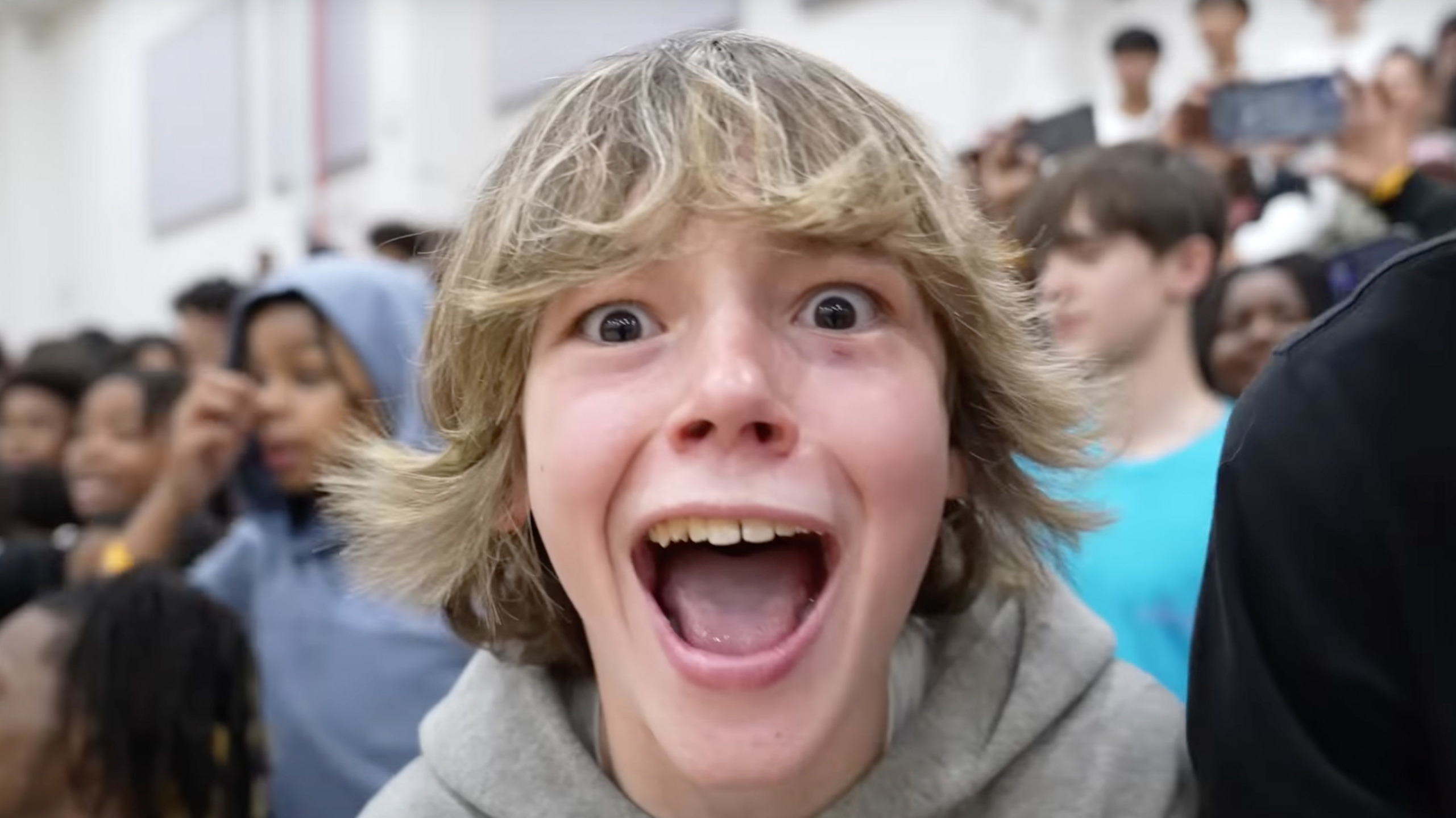 A close-up of a teenage boy with blond shaggy hair looking extremely excited, shouting with his mouth wide open in a crowded hallway.